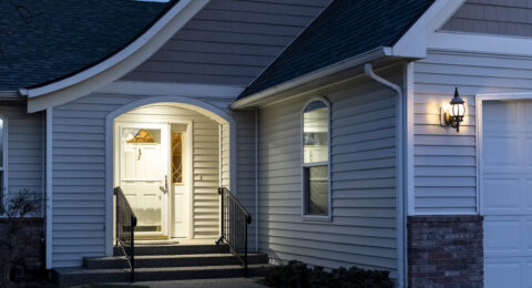 A suburban home’s front porch with a real estate lockbox attached to the railing of the steps is illuminated at evening.